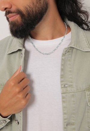 Sølv kædedesignet med sammenkoblede led, båret over en hvid t-shirt, parret med en lysegrøn denimjakke.