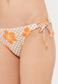 Világos bézs és fehér kockás mintázatú bikini alsó élénk narancssárga virágmintával. Oldalt megkötőkkel rendelkezik a személyre szabható illeszkedés érdekében.