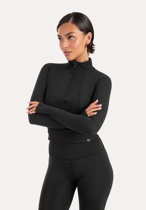 Kvinde med kort mørkt hår iført en tætsiddende sort langærmet lynlåstrøje og sorte leggings, armene krydsede, kigger mod venstre.
