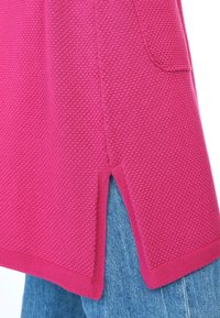 Fuchsia-farbene Oberteil mit Textur und seitlichem Schlitz, kombiniert mit blauer Jeans. Der Stoff hat ein Wabenmuster und einen gerippten Saum.