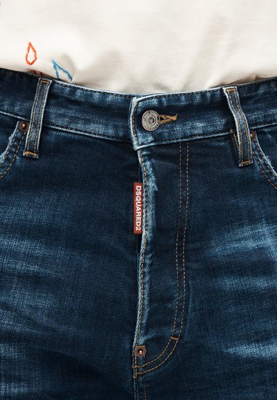 Κοντινό πλάνο μπλε τζιν με ετικέτα DSQUARED2, κλείσιμο με κουμπί και εμφανείς ραφές, φορεμένα με λευκό πουκάμισο μέσα.