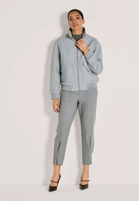 Junge Frau steht vor einer schlichten Wand und trägt eine graue Zip-Jacke, passende cropped Hosen und schwarze spitze High Heels mit Knöchelriemen.