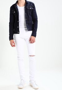 Man med mörkblå denimjacka över vit t-shirt, vita trasiga slim fit-jeans och vita sneakers står mot vit bakgrund.