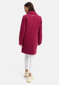 Fuchsia wollen jas met een getailleerde pasvorm, notch-kraag en logodetail op de rug, gecombineerd met witte broeken en beige sneakers.