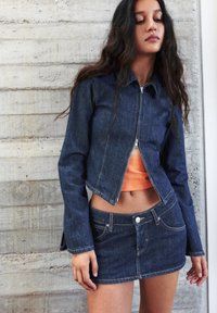Giacca in denim scuro con zip, dal design cropped e maniche lunghe, abbinata a una mini gonna in denim dello stesso colore. Maglietta arancione sotto.