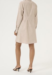 Robe chemise en suede beige avec des manches longues et des plis au dos, atteignant le mi-thigh. Portée avec des sandales à talons aiguilles noires.