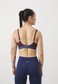 Un reggiseno sportivo bordeaux con spalline regolabili e una fascia blu, abbinato a leggings navy a vita alta. Tessuto liscio con una leggera texture.