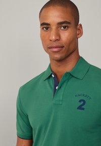 Grønn polo-skjorte med marineblå krage og brodert "HACKETT 2" på venstre bryst. Kort ermer, teksturert stoff og klassisk passform.