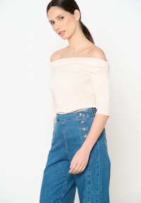 Off-white bezplecu tops ar drapējumiem, kombinēts ar augsto jostasvietu džinsa biksēm ar zelta akcentiem un sānu pogām.