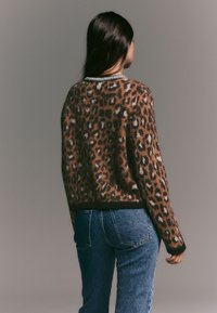 Leopardikuvioinen neule ruskeissa sävyissä, pehmeällä tekstuurilla, pyöreällä kaula-aukollla jossa musta reunus, yhdistetty sinisiin farkkuihin.