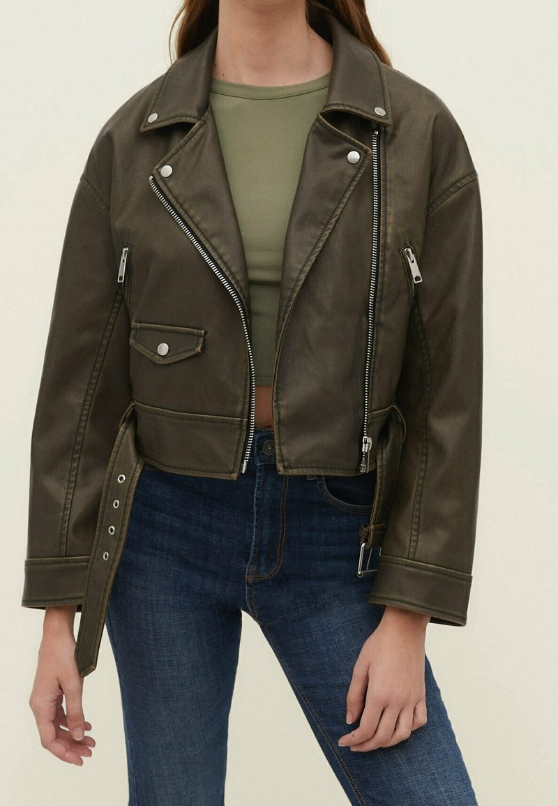 Giacca biker cropped in finta pelle verde oliva con zip e bottoni a pressione in argento, dotata di colletto a risvolto e tasche laterali.