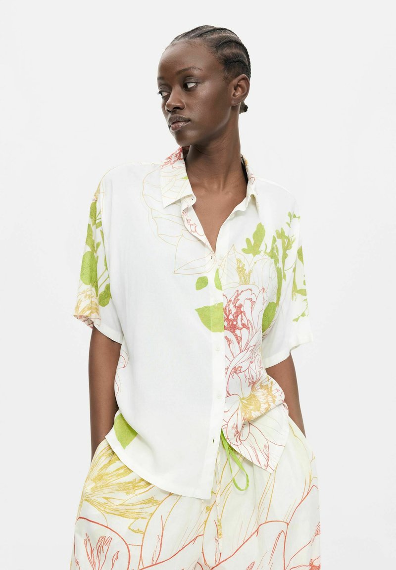 Femme aux cheveux tressés portant une chemise blanche ample et un short assorti avec des motifs floraux verts et orange, debout mains dans les poches.