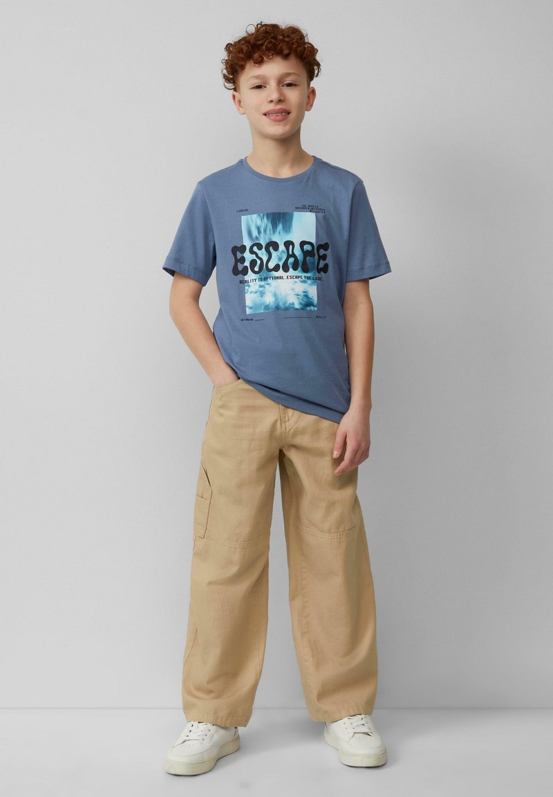 Himmelblaues Baumwoll-T-Shirt mit „ESCAPE“-Grafik in Schwarz und Blau; kombiniert mit beigen Cargohosen und weißen Sneakers.