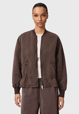 Kobieta ubrana w brązową kurtkę bomberkę na suwak z elastycznym pasem i mankietami, nałożoną na biały top, stojąca na tle jednolitego jasnoszarego tła.