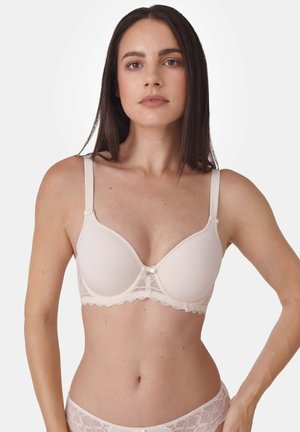 Frau mit langen dunklen Haaren, die einen passenden hellbeigen Spitzen-BH und Spitzen-Slips trägt, steht vor einem schlichten weißen Hintergrund.