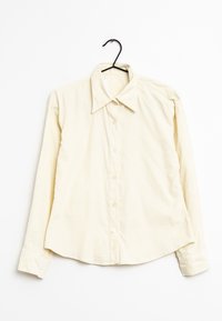 Hellgelbes Langarmhemd mit spitzem Kragen, vorderen Knöpfen und weicher Textur, hängt an einem schwarzen Haken vor einem weißen Hintergrund.