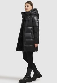 Sort vinterjakke med en blank finish, der har hætte, lynlås lukning og quiltet design. Kombineret med sorte snørebåndsstøvler og leggings.