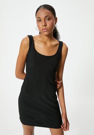 Giovane donna con capelli ricci in abito nero senza maniche, in piedi davanti a uno sfondo chiaro e uniforme, guarda avanti.