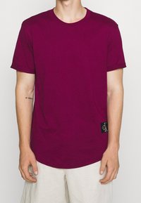 Hombre vistiendo una camiseta de manga corta en magenta oscuro y liso, con una pequeña etiqueta negra de Calvin Klein cerca del dobladillo, combinada con pantalones cortos claros.