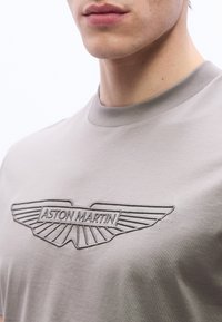 Κοντινό πλάνο ενός ατόμου που φοράει ανοιχτό γκρι πουκάμισο με το κεντητό μαύρο λογότυπ ο της Aston Martin στο στήθος.