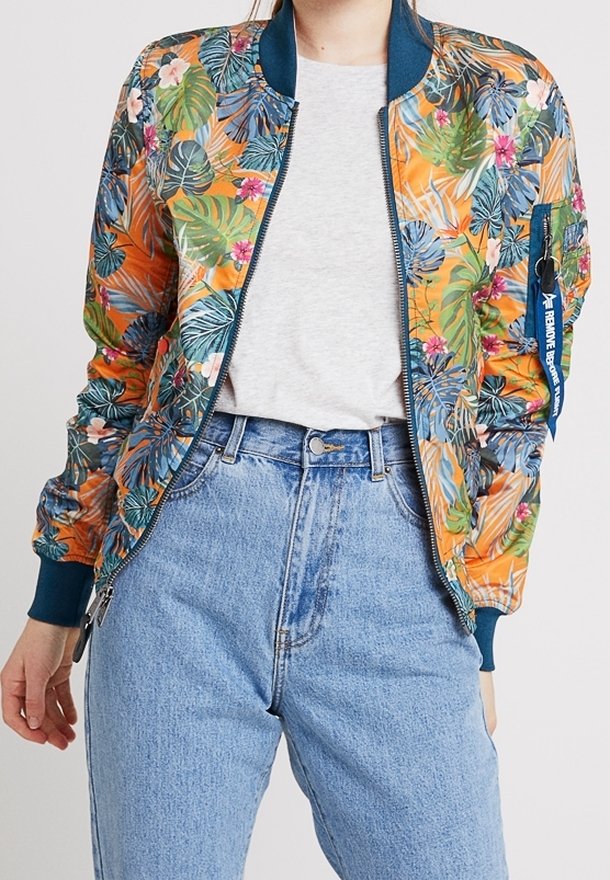 Femme portant une veste bomber florale vibrante avec des feuilles vertes, bleues et roses sur un t-shirt blanc et un jean taille haute bleu clair.