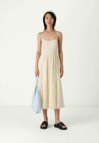 Rochie maxi fără mâneci, de culoare crem, cu țesătură texturată, talie strânsă și formă în A. Asortată cu o geantă bleu deschis și sandale negre.