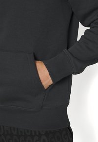Sweatshirt gris foncé avec une poche kangourou, des poignets côtelés et une finition texturée. Le matériau est doux avec une légère élasticité.