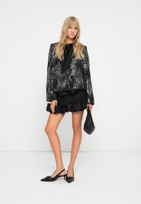 Zwarte paillettenblazer met gestructureerde schouders, gedragen over een zwarte top en een gerimpelde rok. Het model houdt een zwarte handtas vast en draagt flats.