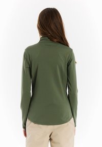 Langsleeve olijfgroen sporttop met een opstaande kraag, gladde stof en een licht gebogen zoom, gecombineerd met lichtbeige broeken.