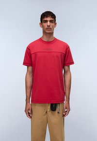 Κόκκινο κοντομάνικο μπλουζάκι με στρογγυλή λαιμόκοψη, με οριζόντια ραφή και κεντημένο λογότυπο. Φοριέται με χακί παντελόνι και μικρό αξεσουάρ.