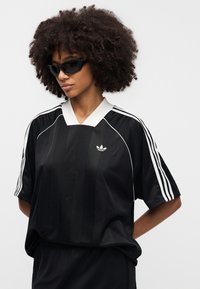 Μαύρο αθλητικό φανέλα με λευκές ριγέ λεπτομέρειες και λευκό γιακά. Διαθέτει μικρό λογότυπο του εμπορικού σήματος στο στήθος. Μαλακό, λείο ύφασμα.