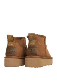 Botas de ante marrón hasta el tobillo con una superficie texturizada, que presentan un parche con el logo y una tira de tela para facilitar el calzado, además de una suela de goma gruesa y resistente.