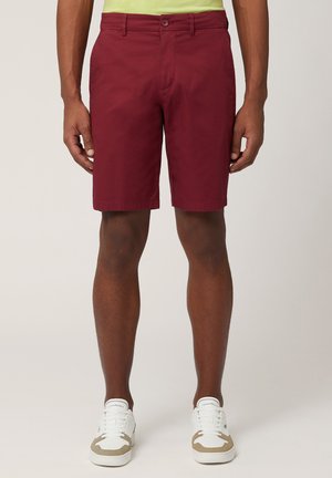 Bordeauxkleurige stofshorts met een platte voorkant, ceintuurlussen en zijzakken; gecombineerd met witte sneakers met beige accenten.