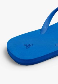 Tongs bleues en caoutchouc, dotées d'une semelle Texturée et d'une lanière lisse. Logo embossé sur le côté. Bords légèrement arrondis.