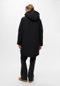 Schwarze Daunenjacke mit Kapuze, elastischen Bündchen und einem längeren Schnitt. Kombiniert mit dunklen Jeans und hellbraunen Schuhen.