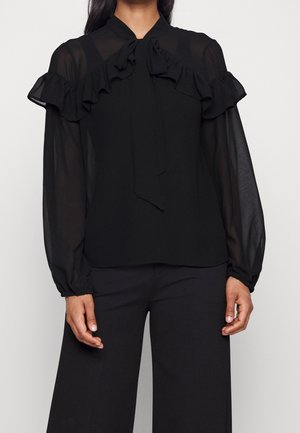 Woman wearing a black blouse with sheer long sleeves, ruffled details on shoulders, and a tied bow at the collar, paired with black pants.
