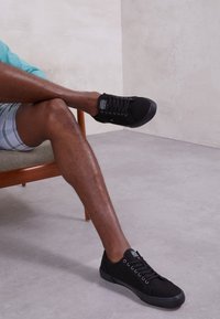 Baskets en toile noire avec bout arrondi, semelle plate et design à lacets. Portées avec un short à carreaux. Texture lisse, branding minimal visible.