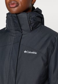 Zwarte Columbia-jack met hoge kraag, voorzien van een rits, verstelbare capuchon en wit logo op de bovenste linkerkant. Glad textuur.