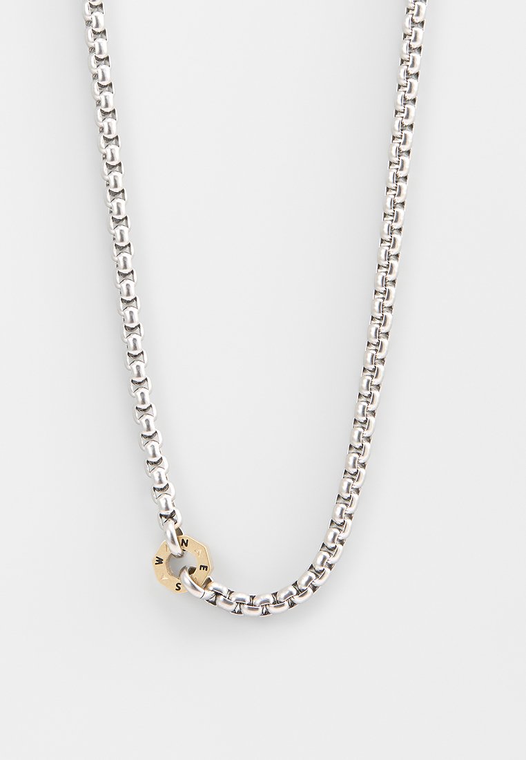 Silberne Metallkette mit glatter Oberfläche; bietet einen runden goldenen Akzent, der mit Buchstaben graviert ist, was Textur und Kontrast hinzufügt.