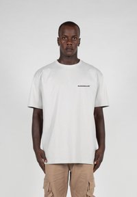Light grey cotton t-shirt with a relaxed fit, short sleeves, and a logo printed in black on the left chest. Plain design, soft texture.
