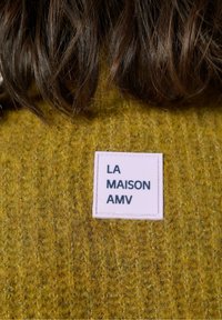 Gorčično rumena pletena jopica z teksturiranim zaključkom, ki ima kvadratno rožnato oznako z besedilom "LA MAISON AMV" v črnem tisku.