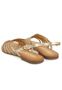 Sandales plates tressées en or avec une sangle et une boucle, dotées d'une semelle lisse marron et d'une semelle intérieure texturée arborant le nom de la marque imprimé.