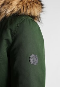 Grøn parka-jakke med en brun kunstpelshætte. Har et sort mærke på ærmet med prægede brandlogo og året 1961.