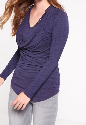 Femme portant un haut bleu foncé à manches longues froncé avec des boutons près de l’encolure et un pantalon gris clair, sur un fond uni.