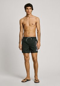 Sorte svømmebukser med hvidt logotekst, elastisk talje og snøre. Båret med sorte flip-flops, model står mod en ensfarvet baggrund.