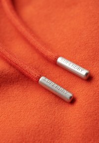 La tela de la sudadera naranja presenta dos cordones con terminales metálicos plateados, grabados con "SUPERDRY." Primer plano de la textura y detalles del color.