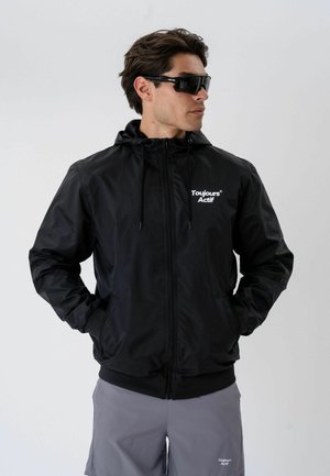 Homme portant une veste à capuche noire zippée et un short gris, les mains dans les poches, avec des lunettes de soleil enveloppantes noires, regardant sur le côté.