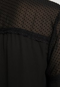 Schwarze Bluse mit einem durchsichtigen gepunkteten Overlay, Spitzenbesatz am Kragen und einem glatten undurchsichtigen Stoffkörper. Texturierte schwarze Punkte sind sichtbar.