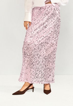 Nő padlót érő rózsaszín flitteres szoknyában, fehér blúzban és barna, alacsony sarkú slingback cipőben, egy egyszínű háttér előtt áll.