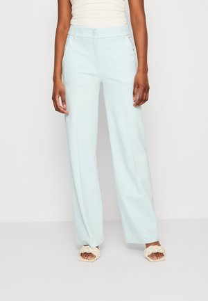 Woman wearing light blue wide-leg trousers and cream braided open-toe sandals standing against a plain wall.